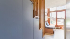 A modern room with a grey wardrobe, a wooden slatted partition, a bed with a grey mattress, and a large window providing natural light. - 300m from resort center, 1st floor, view valley, balcony, tv, 37m², Les Arcs (Les Arcs)