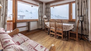 A cozy room with a wooden floor, a sofa, a dining table with chairs, and large windows offering a snowy mountain view. - 20m from ski slopes, 5th floor, balcony, tv, ski locker, 50m², La Plagne (La Plagne)