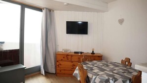 A room with a large window, a flat-screen TV mounted on the wall, a wooden dresser, a table with a checkered tablecloth, and a heart-shaped decoration on the wall. - 15m from ski slopes, 6th floor, balcony, 23m², Les Orres (Les Orres)