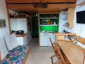 A compact kitchen with a microwave, stove, and sink. There is a wooden dining table with chairs and a sofa with a colorful cushion.