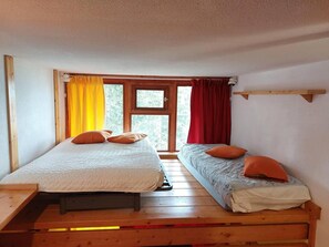 A room with two beds, a window with curtains, and a wooden beam shelf.