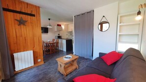 Una sala de estar con un sofá gris oscuro, cojines rojos, una mesa de centro de madera y una cocina al fondo.