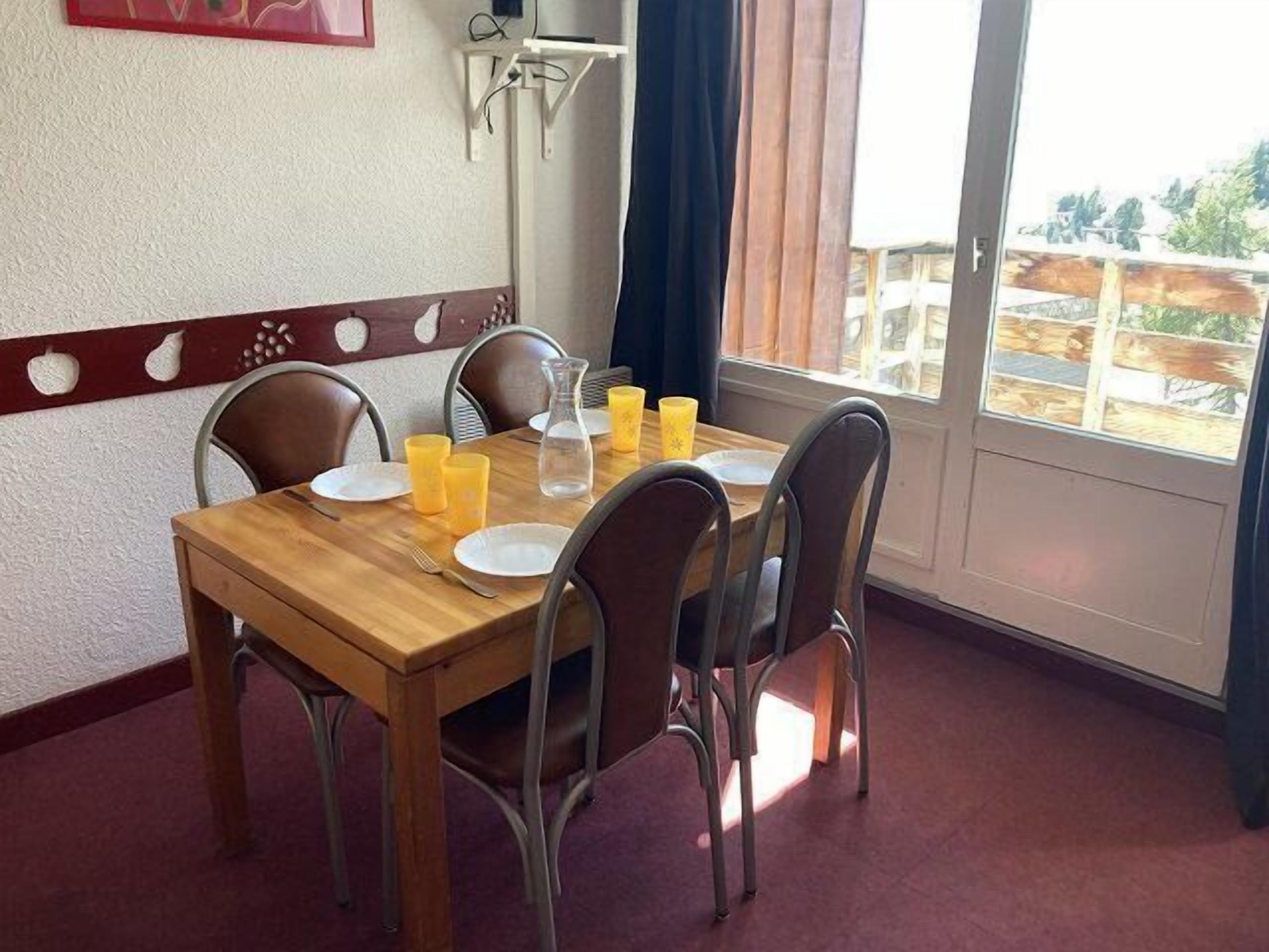 A dining table set with plates, glasses, and a pitcher, surrounded by chairs, with a window and curtains in the background.