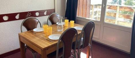 A dining table set with plates, glasses, and a pitcher, surrounded by chairs, with a window and curtains in the background.