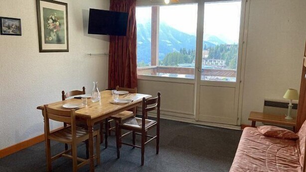 Una stanza con un tavolo da pranzo in legno, due sedie, un televisore a parete, una finestra con vista sulle montagne e un letto con coperta rosa.