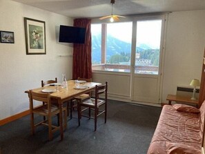 A room with a wooden dining table, two chairs, a television on the wall, a window with a view of mountains, and a bed with a pink blanket. - 50m from ski slopes, 5th floor, tv, 25m², Orcières Merlette 1850 (Orcières)