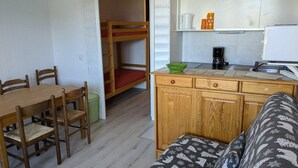 Ein kompakter Raum mit Holztisch und Stühlen, einem Etagenbett, einer Küchenzeile mit Kaffeemaschine und einem Sofa mit gemusterten Kissen.