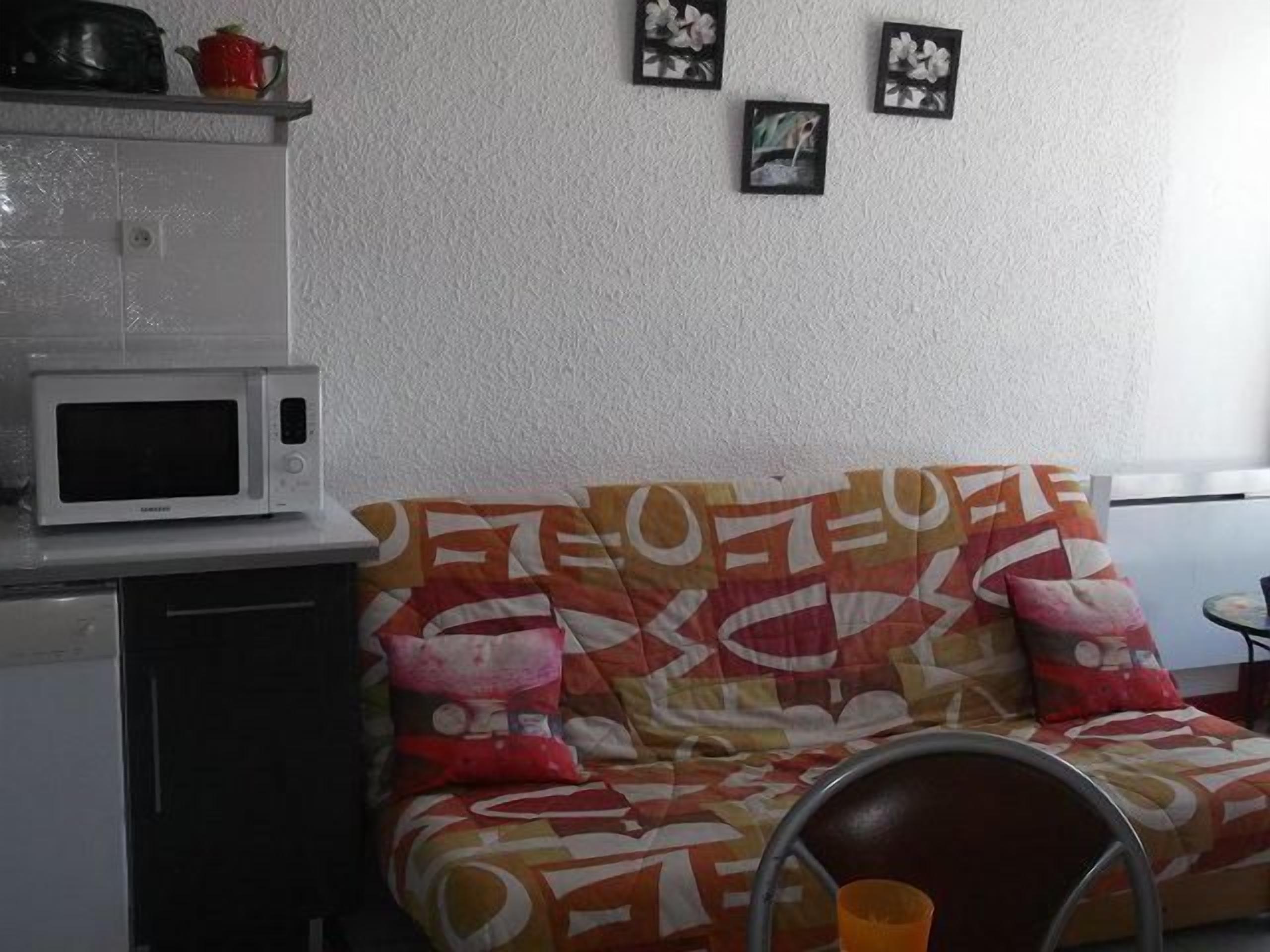 A kitchenette with a microwave, a small refrigerator, and a shelf with kitchenware. There is a colorful sofa with patterned cushions and a metal chair.