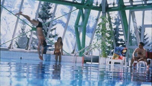 An indoor swimming pool with a glass ceiling, a diving board, and lounge chairs.