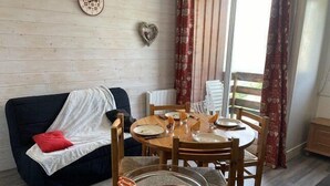 Ein gemütliches Wohnzimmer mit Essbereich. Ein schwarzes Sofa mit rotem Kissen und weißer Decke steht an der Wand. Ein Holztisch mit Tellern, Besteck und einer Schüssel mit roter Füllung ist gedeckt.