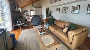 A cozy living room with a wooden ceiling, a brown leather sofa, a coffee table, and a stone fireplace. - The Sauna Cottage on Keuka Lake - Wine Trail Retreat (Keuka Park)