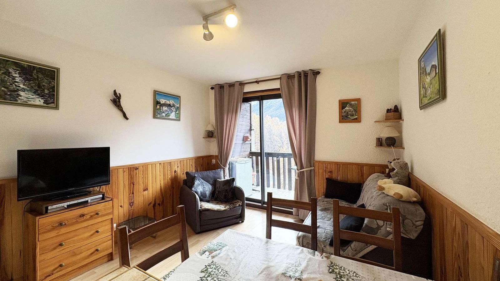 A cozy living room with a television, wooden furniture, and a balcony with curtains.