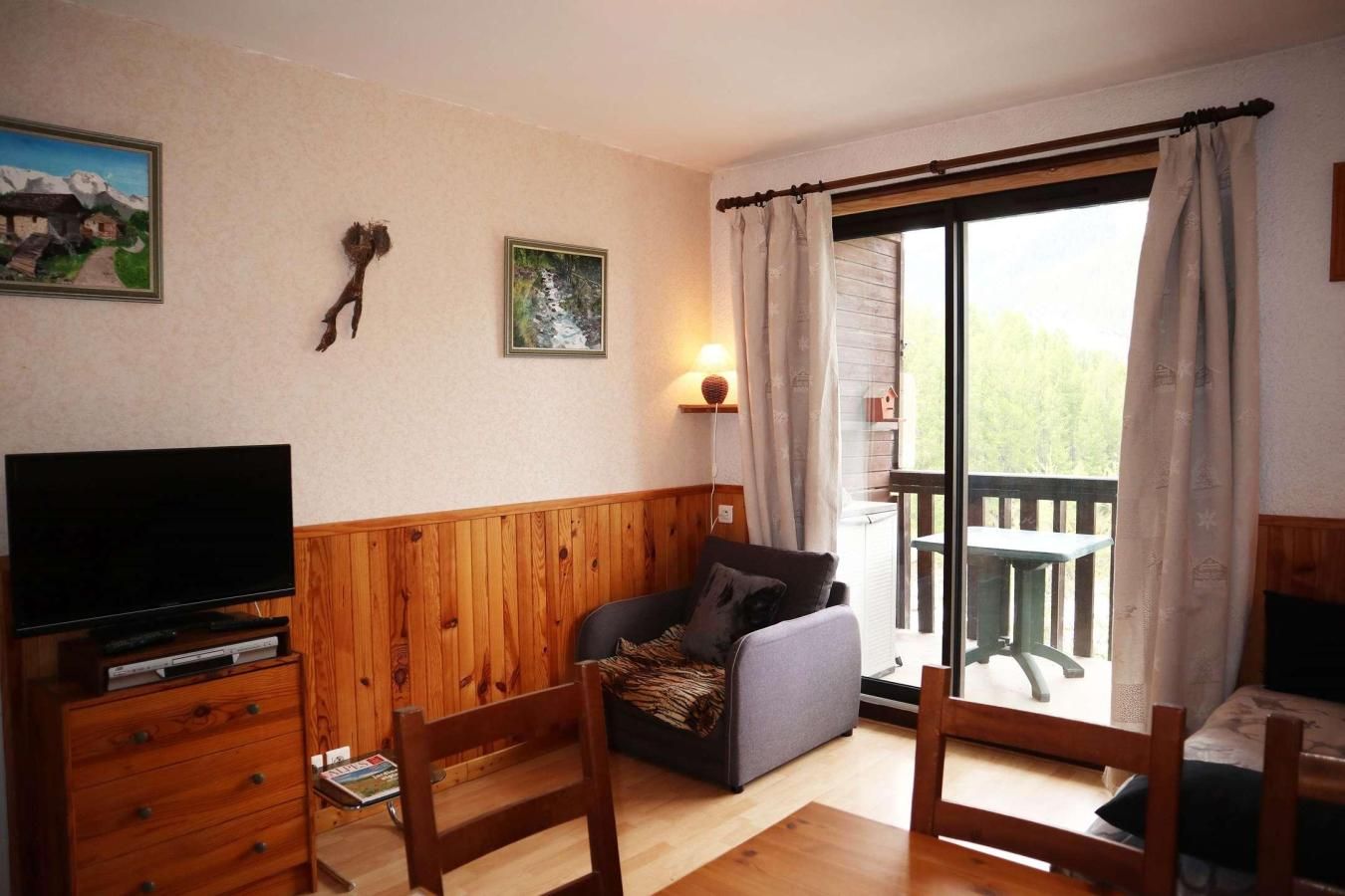 A room with a television, wooden furniture, a sofa, and a balcony with a table and chairs.