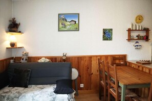 Une pièce confortable avec un lit, une table à manger en bois et des chaises. On y trouve une lampe, une horloge et une étagère avec des objets décoratifs.
