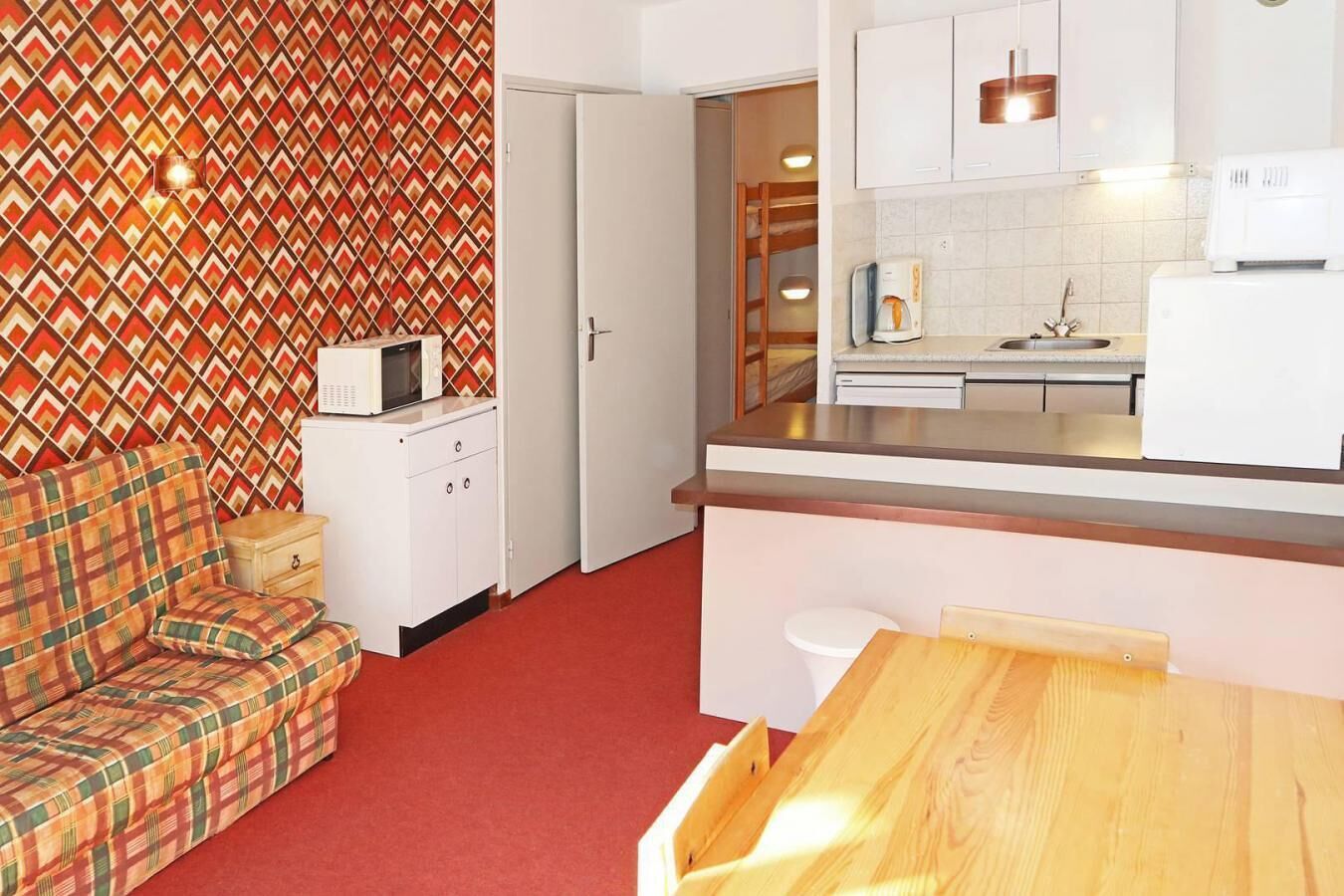 Een kleine keuken met witte kastjes, een magnetron en een koffiezetapparaat. Er is een houten tafel en een ruitjesbank in een woonhoekje.