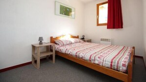 A single bed with a wooden frame, a bedside table with a lamp, a wall-mounted picture, and a window with a red curtain. - 150m from ski slopes, 4th floor, view mountain, balcony, parking, ski locker, 36m², Valfréjus (Valfréjus)