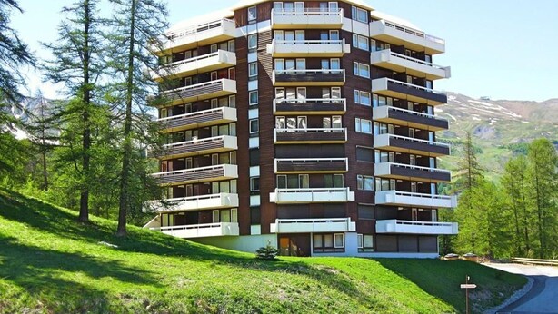 Un edificio de varios pisos con balcones, rodeado de árboles y un paisaje montañoso.
