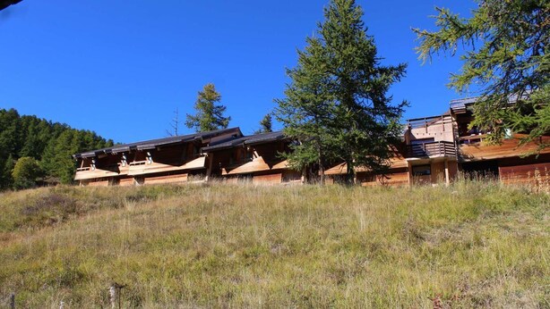 Une cabane en bois à plusieurs niveaux, entourée d’un champ verdoyant et d’arbres.