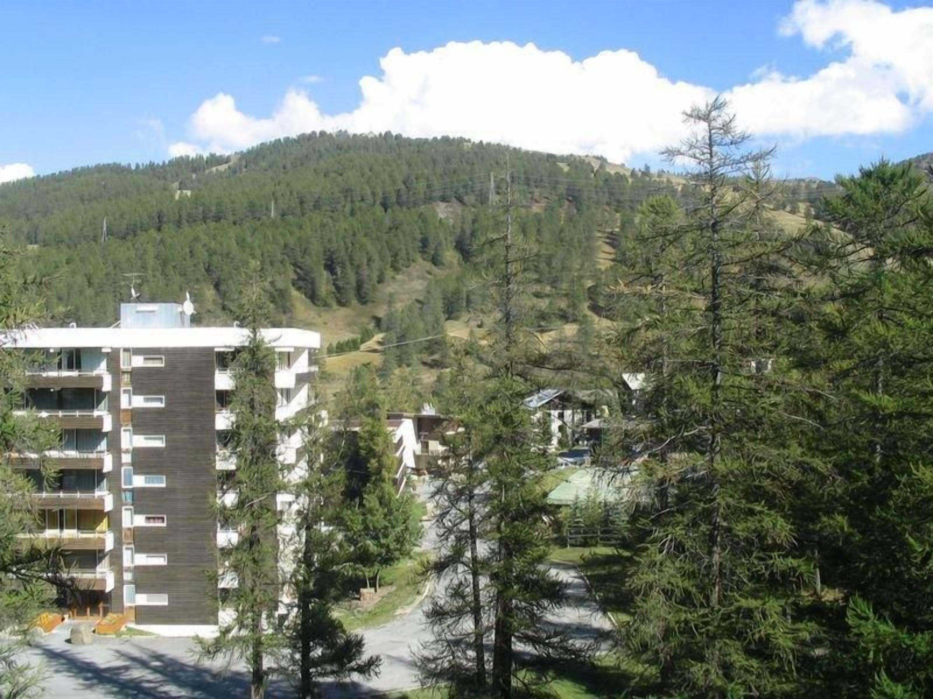 A multi-story building with balconies, surrounded by trees and a hilly landscape.