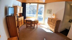 A compact room with a wooden cabinet, a television, a dining table with chairs, a dresser, and a window with a view of trees.
