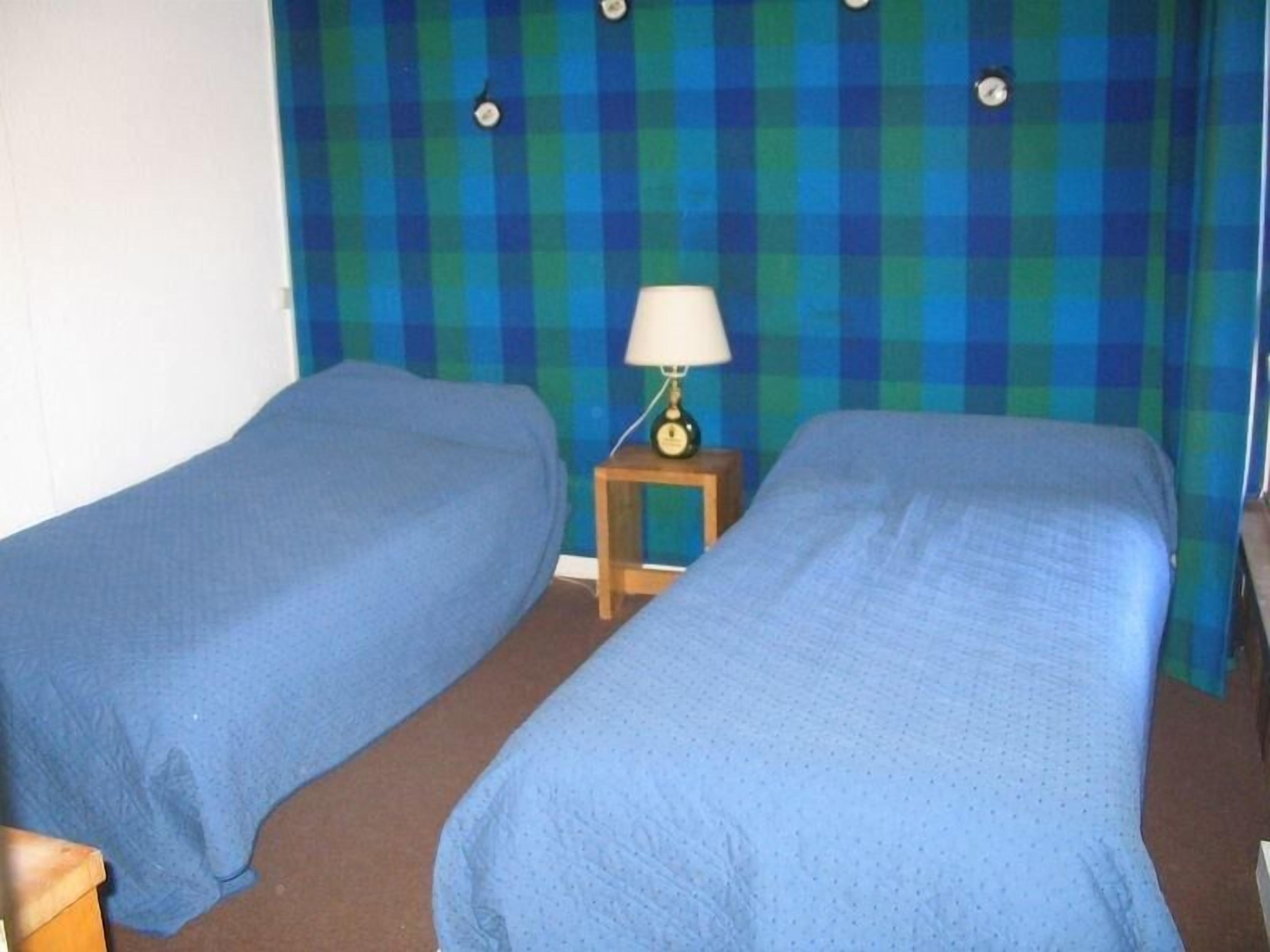Une chambre avec deux lits simples, une petite table de chevet en bois avec une lampe et un mur à carreaux.