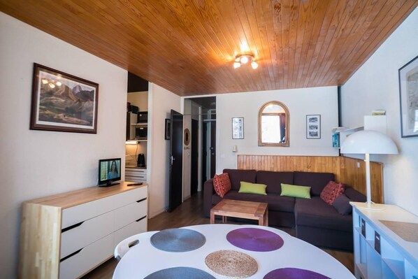 A cozy living room with a wooden ceiling, a sofa with cushions, a coffee table, and a TV on a wooden cabinet.