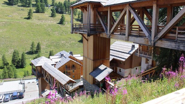 A wooden chalet with a balcony and multiple levels, surrounded by a mountainous landscape.