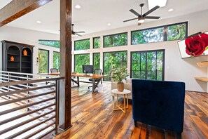 A spacious room with a wooden floor, a desk, a blue chair, and large windows offering a view of greenery.