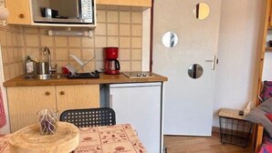 Ett kompakt kök med mikrovågsugn, kaffebryggare och ett litet kylskåp. Där finns ett matbord med en mönstrad duk och en stol.