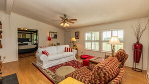 A living room with a white sofa, patterned armchair, and a red armchair. There is a ceiling fan, a red lamp, and a vase on a side table. - Rural Retreat with Private Pond, ATV Trails and Event Space (Buffalo)