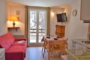 A cozy room with a red sofa, a dining table with chairs, a TV mounted on the wall, and a window with a view of trees.