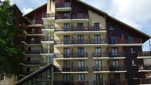 Un hotel de varios pisos con balcones en cada planta, entrada de cristal y coches estacionados frente a él.