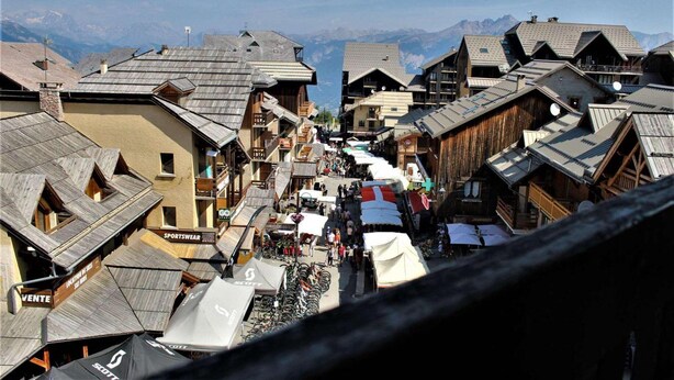 Une rue étroite bordée de bâtiments en bois, dont certains sont dotés de balcons, avec une chaîne de montagnes en toile de fond.