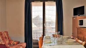 Un coin repas avec une table dressée pour quatre personnes, un téléviseur posé sur un meuble et une vue sur un bâtiment à travers des rideaux à motifs.
