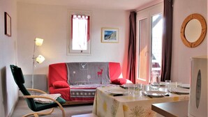 Una habitación acogedora con una mesa de comedor dispuesta para dos personas, una mecedora, una mesita auxiliar y una cama con manta roja y gris.