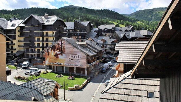 Un pueblo con edificios de madera tradicionales, un edificio central con un gran letrero que anuncia un complejo de esquí, y montañas al fondo.