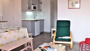 Un espacio habitable compacto con kitchenette, un área de comedor con mesa y banco, un sillón verde y un pequeño sofá rojo.