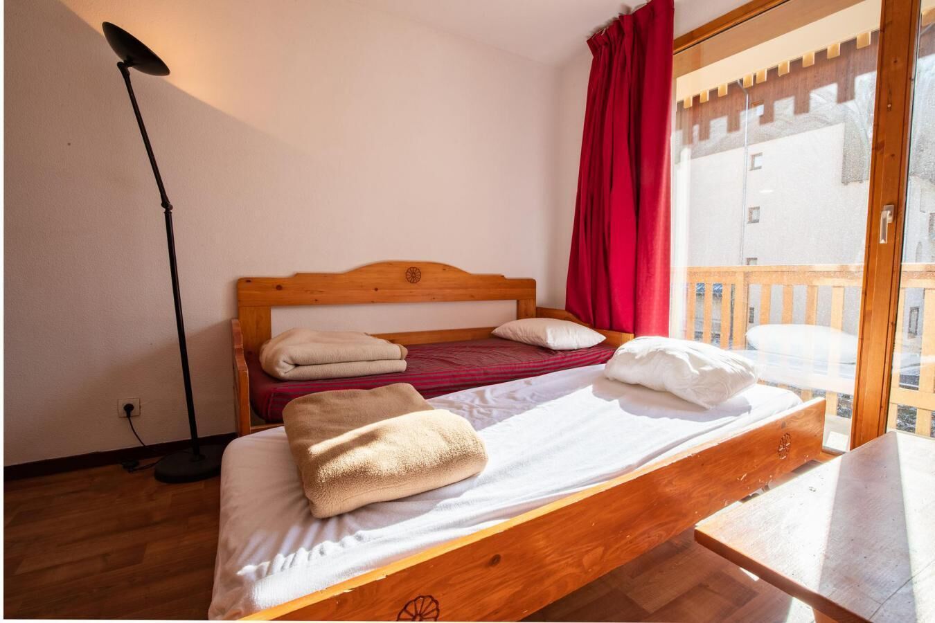 A bedroom with a wooden bed, two pillows, a red curtain, and a view of a wooden deck through a glass door.