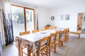 A dining area with a long table and chairs, a window with curtains, and a small cabinet.
