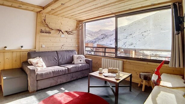 Una habitación acogedora con techo de madera, un sofá gris, una mesita con un reposapiés rojo y un ventanal con vistas a la montaña.