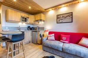 A compact kitchen with wooden cabinets, a microwave, and a coffee machine. There is a red sofa with patterned cushions and a small table with chairs.