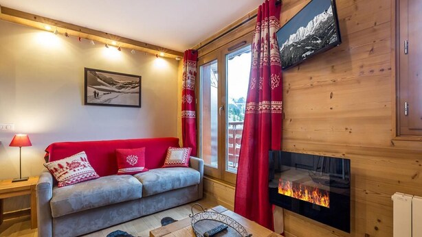 Een gezellige woonkamer met een grijze bank, rode kussens, een houten vloer, een open haard en een aan de muur bevestigde televisie.