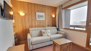 Um quarto aconchegante com sofá, uma mesinha, paredes de madeira, uma TV na parede e uma janela com cortinas.