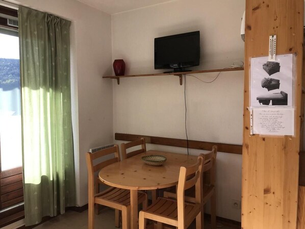 A room with a round wooden table, four chairs, a television mounted on the wall, and a window with green curtains. - 2nd floor, view mountain, balcony, 28m², Villard de Lans (Villard-de-Lans)