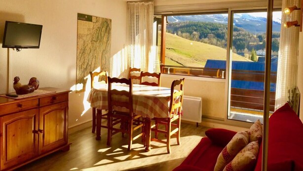 A dining area with a table and chairs, a TV on a stand, a map on the wall, and a view of a mountainous landscape through large windows. - 3rd floor, view mountain, balcony, tv, 33m², Villard de Lans (Villard-de-Lans)