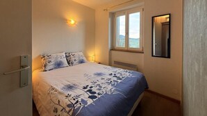 Um quarto com cama, janela, espelho fixado na parede e abajur.