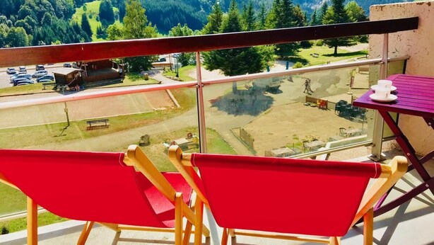 A balcony with red chairs and a table, overlooking a mountainous landscape with a parking area and buildings in the distance.