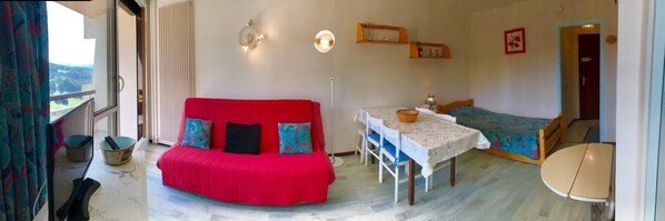 Una habitación acogedora con un sofá rojo, una pequeña mesa de comedor, una cama con colcha de colores y vistas al exterior.