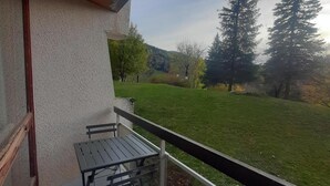 Un balcón con una mesa y sillas, con vista a una zona verde con árboles y una colina al fondo.