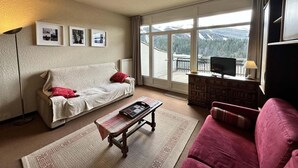 Una sala de estar con sofá, un sillón rojo, una mesa de centro, un televisor sobre un mueble de madera y un ventanal con vistas a la montaña.